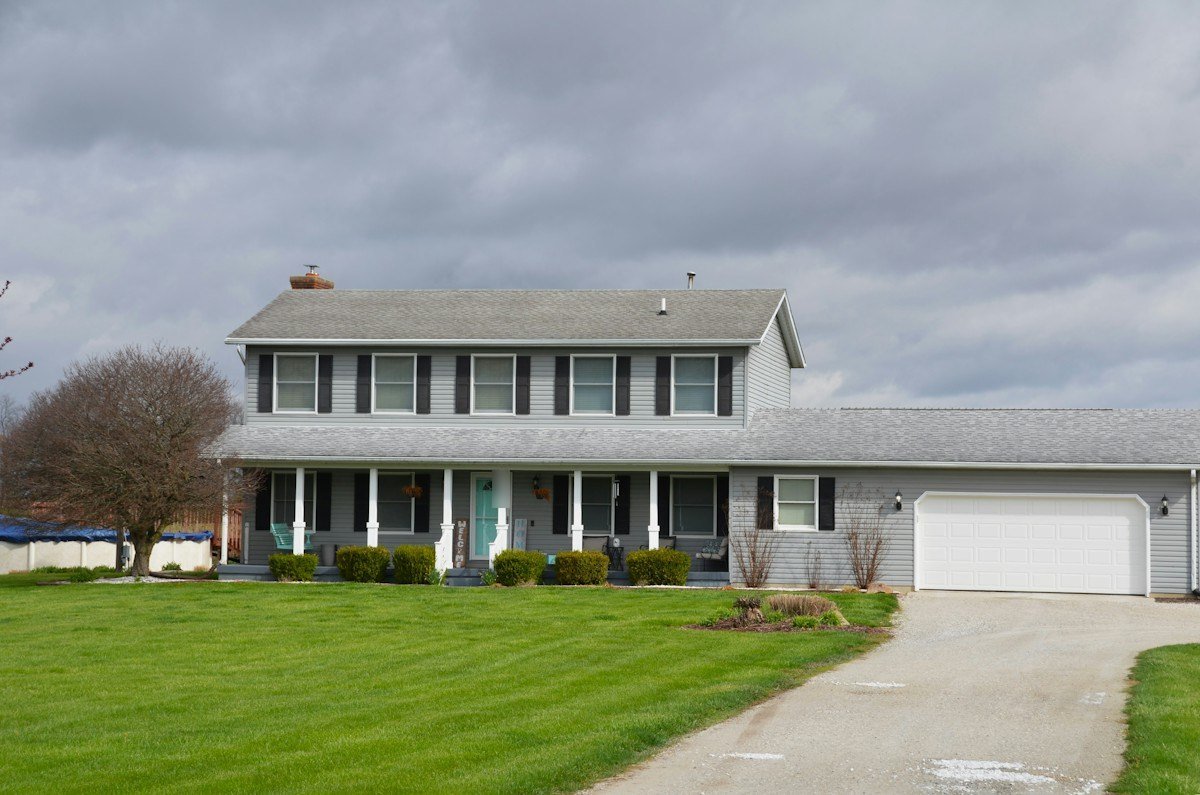 A gray two-story house with a long front porch under a cloudy sky
