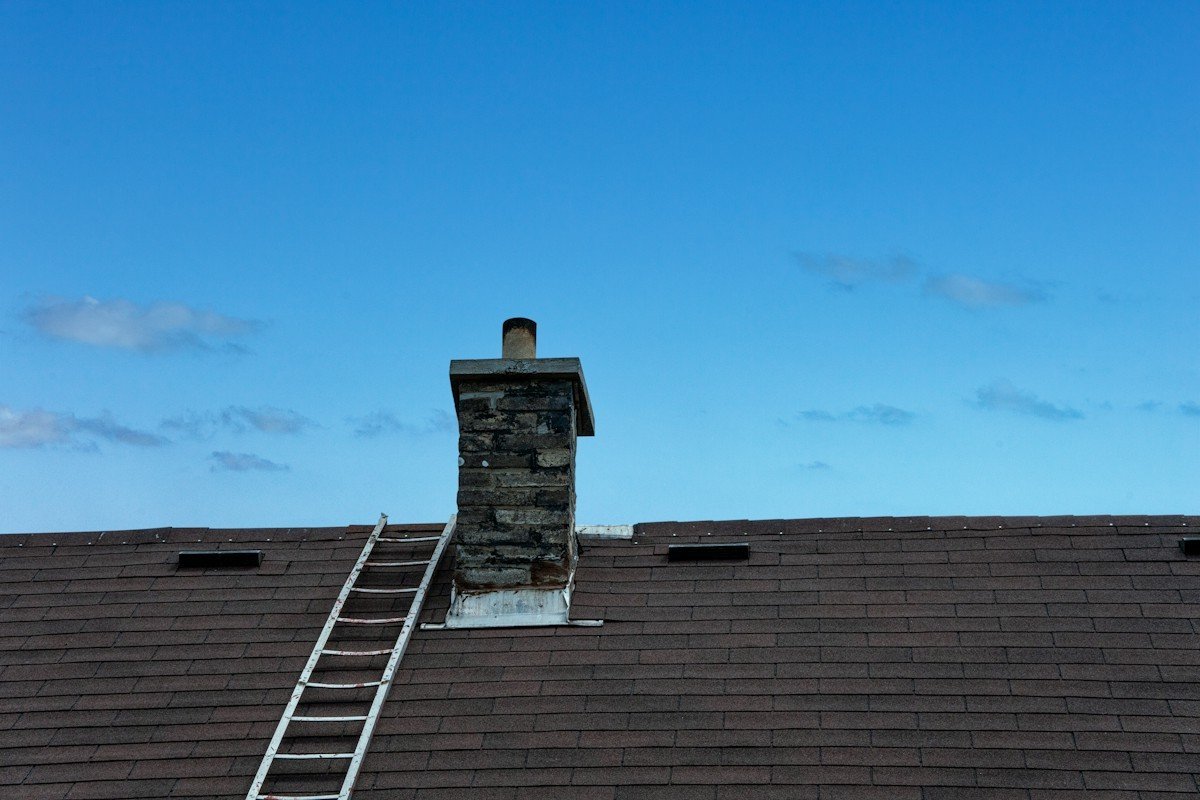 A ladder resting on a brown shingle roof beside a stone chimney