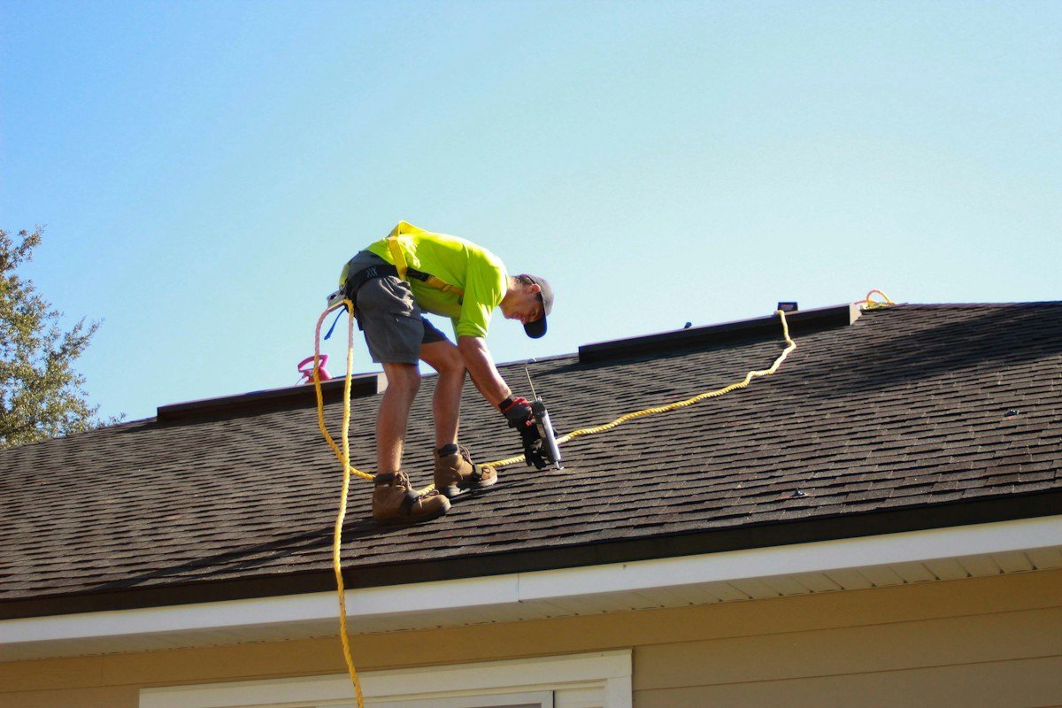 A roofer in a safety harness nailing down shingles with a nail gun