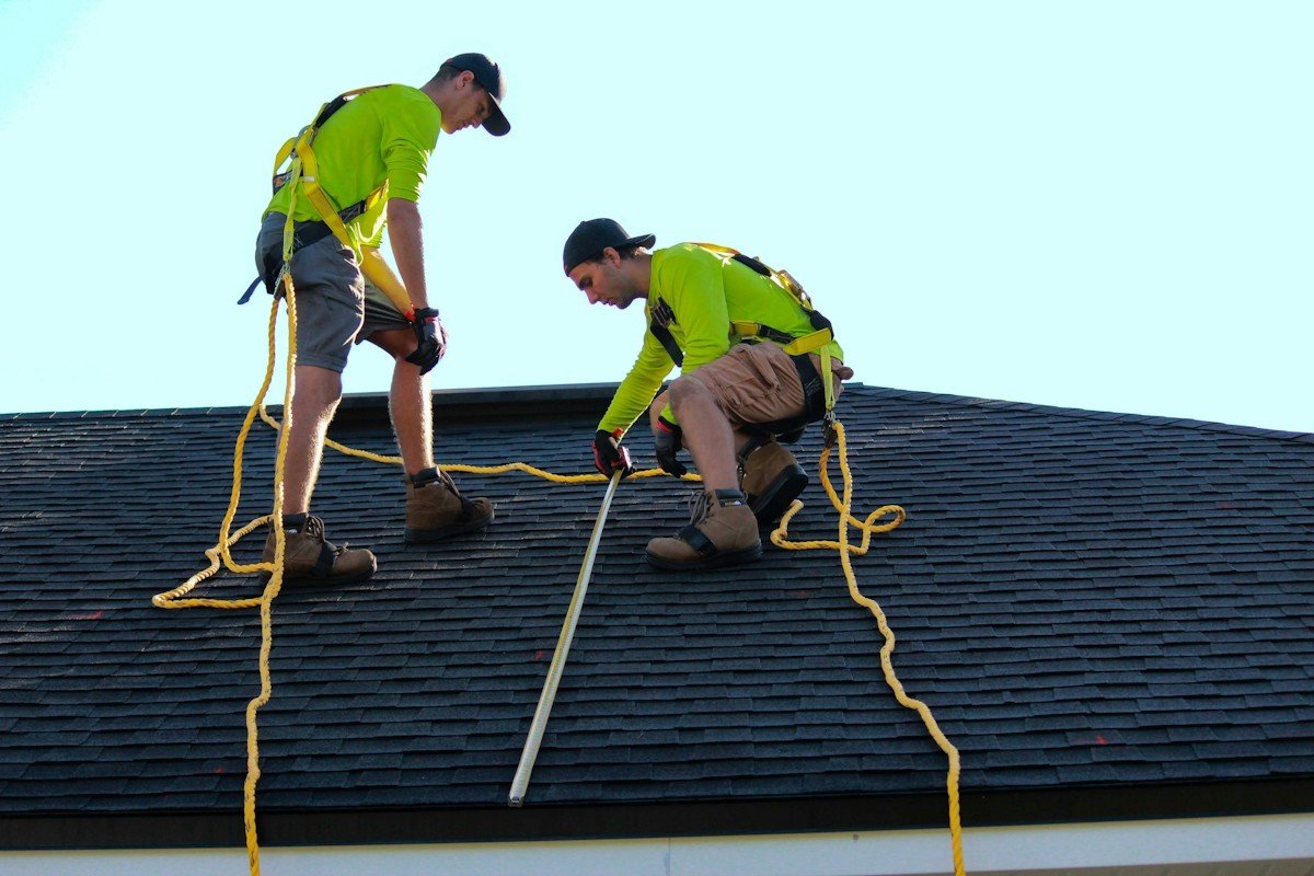 Two roofers in safety harnesses measuring a dark shingle roof